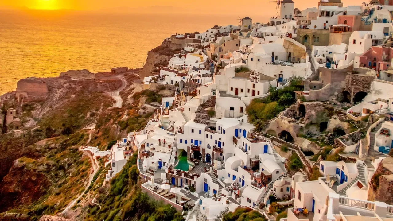 The whitewashed cliffside houses and windmill of Oia glowing at sunset over the Santorini caldera