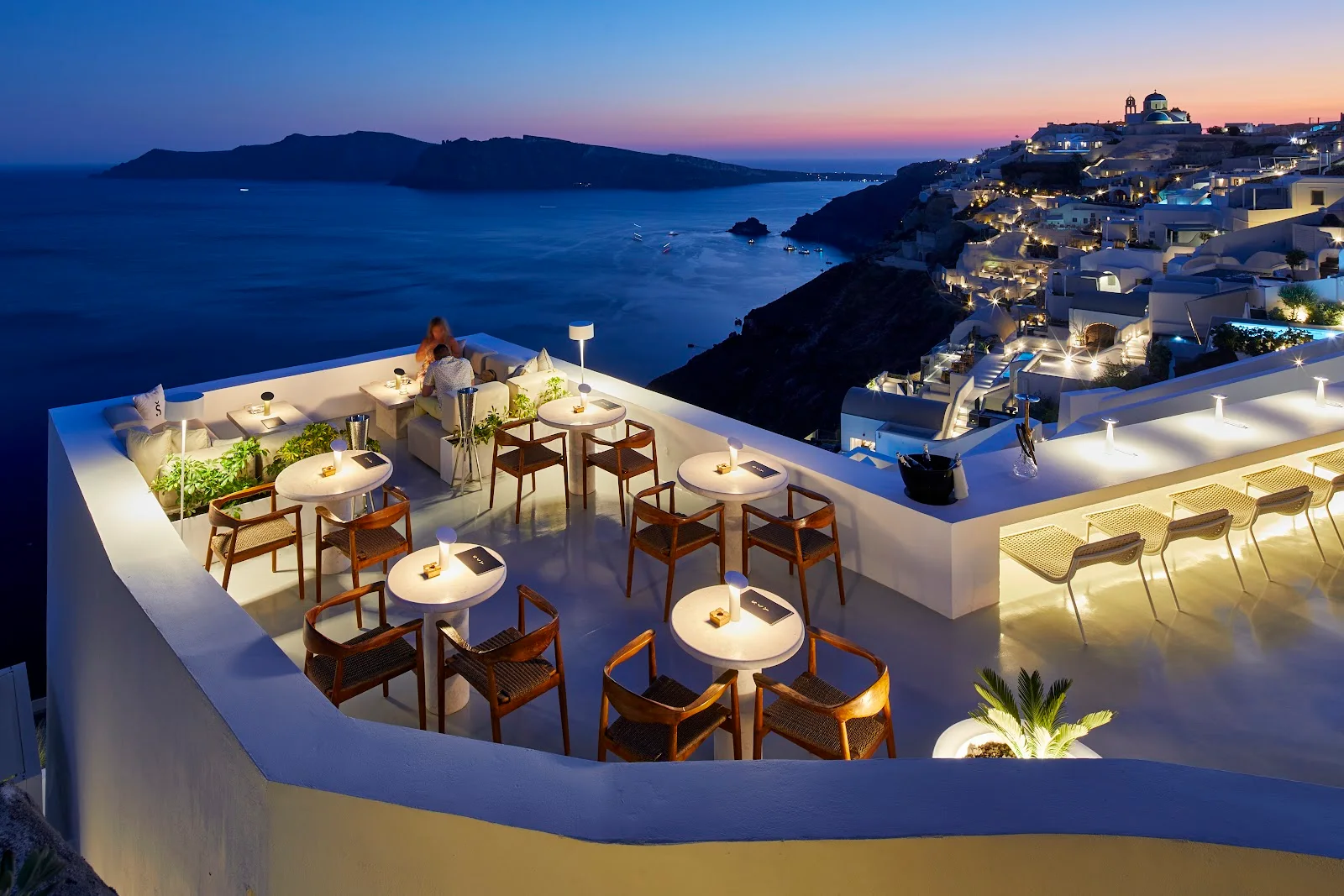The white rooftop terrace with rattan chairs looking over the Oia caldera and sea at dusk, Saint Sky Bar