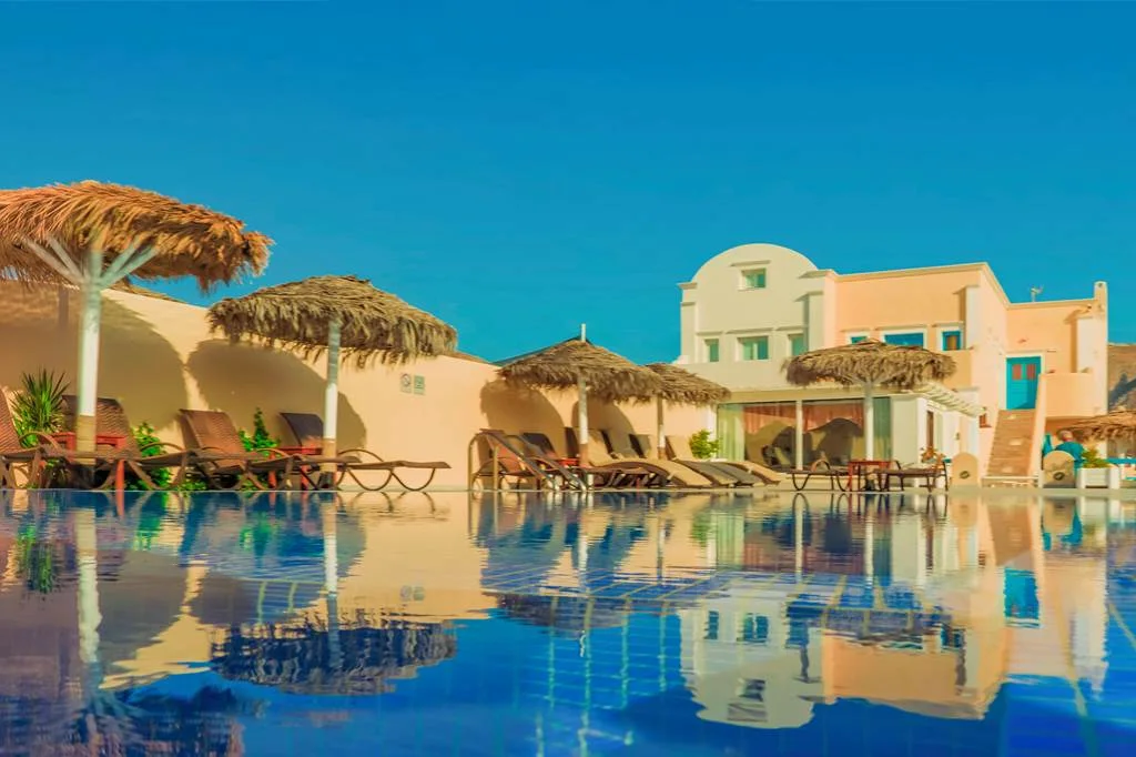 The pool, sun loungers and thatched umbrellas of Perissa Bay Hotel below its Cycladic buildings in Perissa, Santorini