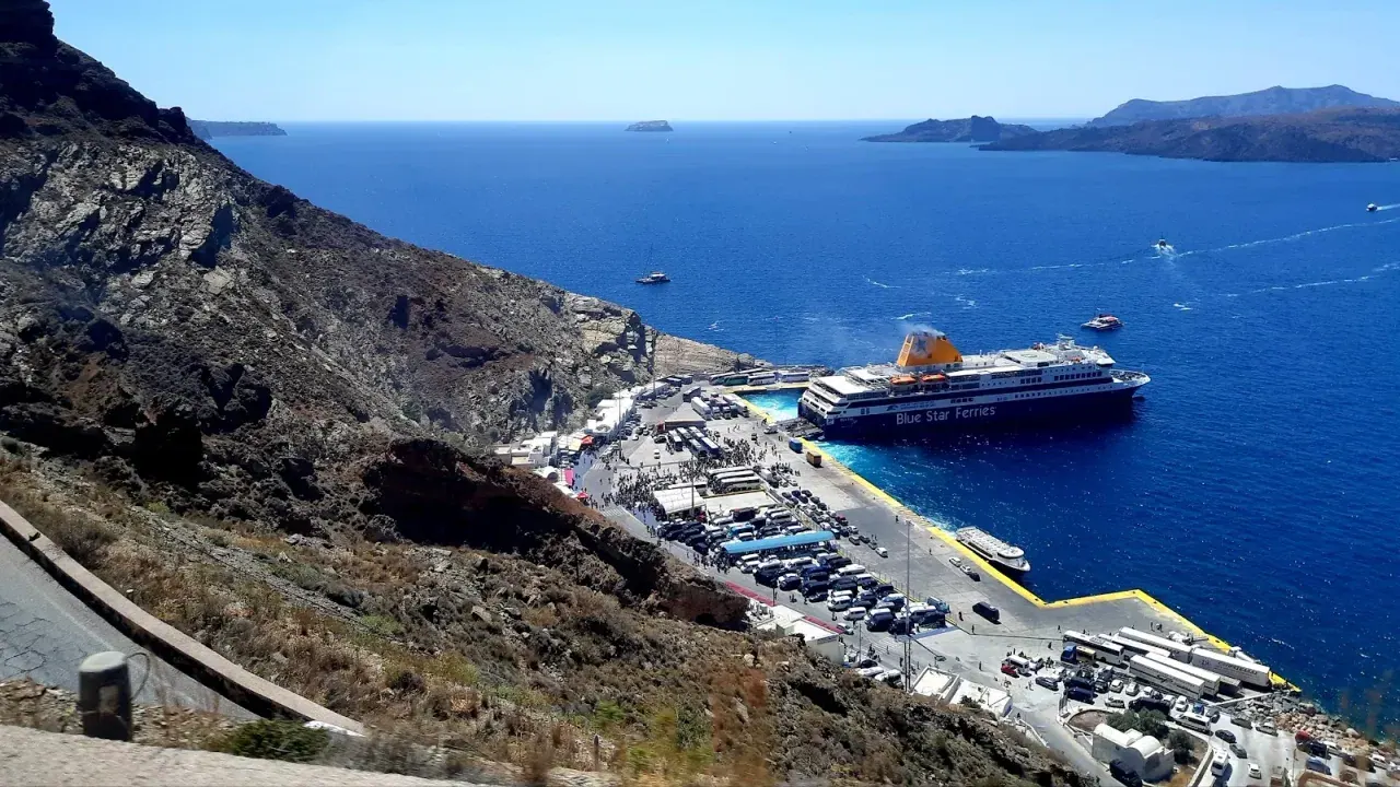 A Blue Star ferry docked at Athinios port below the switchback cliff road, with the Santorini caldera beyond
