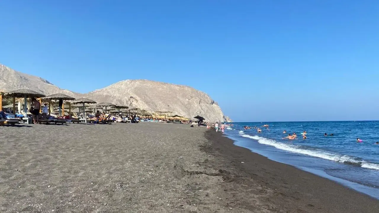 Straw umbrellas along the dark volcanic sand of Perivolos beach with swimmers in the sea below Mesa Vouno, Santorini