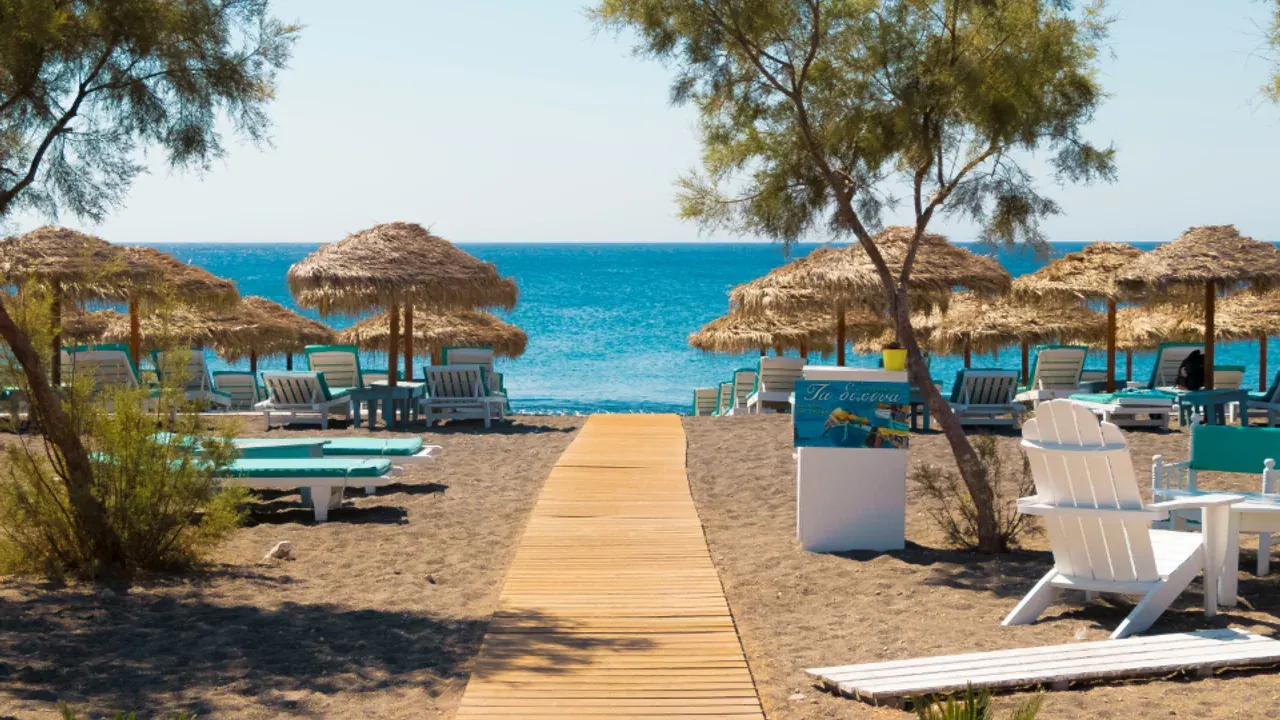 Straw parasols and sunbeds on a black-sand Santorini beach, a boardwalk leading to the sea