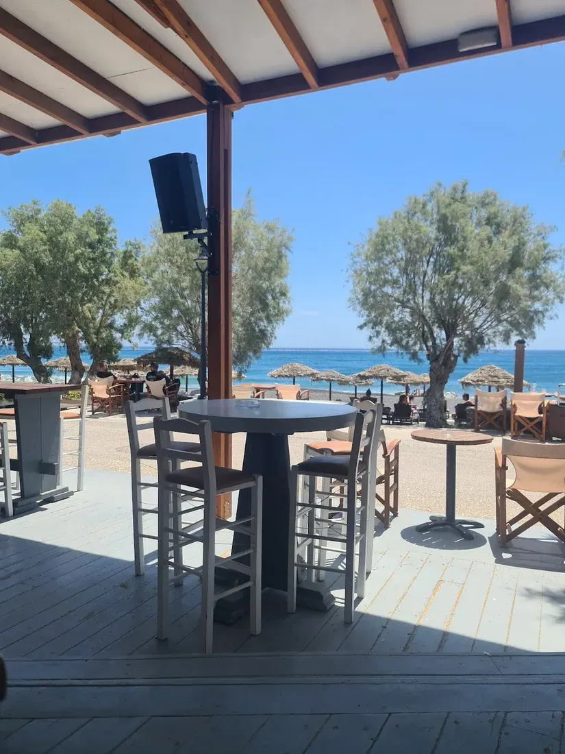 Covered wooden deck with bar stools looking out to thatched umbrellas and the sea at The Beach Bar, Perissa