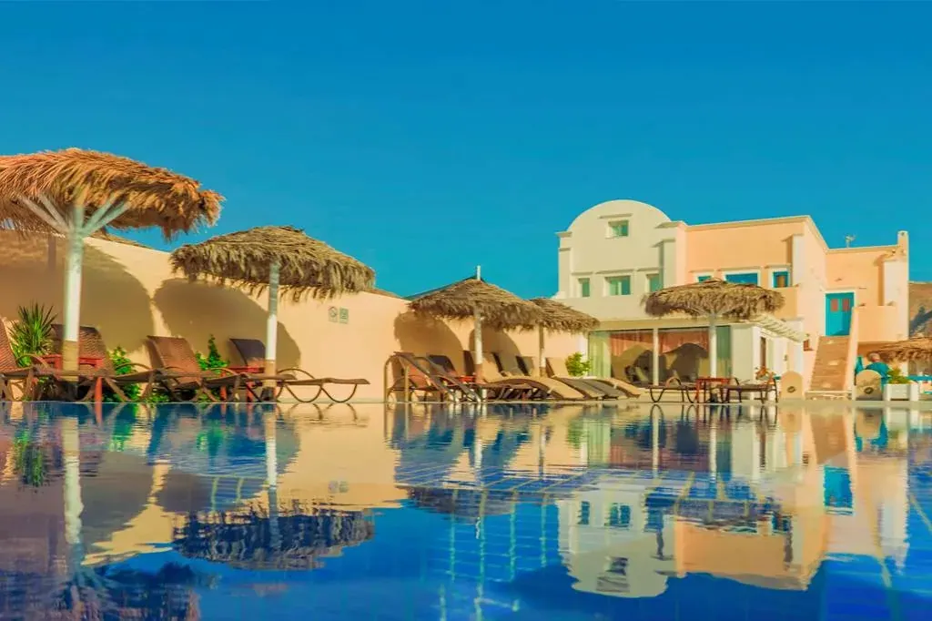 The pool, sun loungers and thatched umbrellas of Perissa Bay Hotel below its Cycladic buildings in Perissa, Santorini