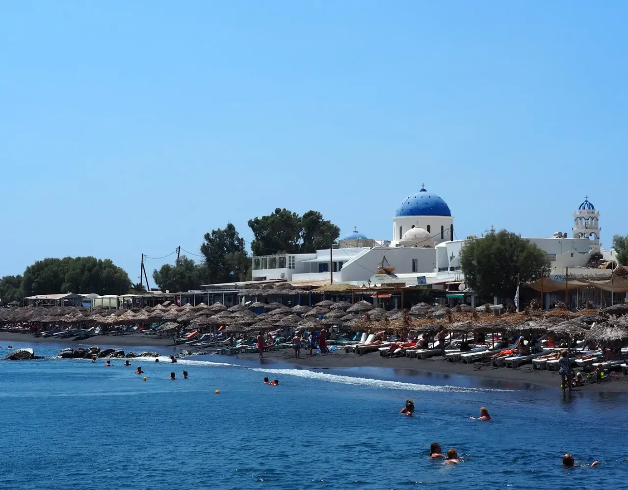 The black volcanic sand of Perissa Beach lined with straw umbrellas below the Mesa Vouno rock and a blue-domed church
