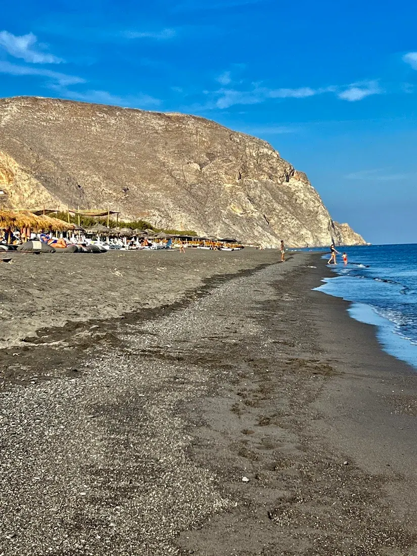 The black volcanic sand of Perivolos beach stretching below Mesa Vouno, with sunbeds and the Aegean