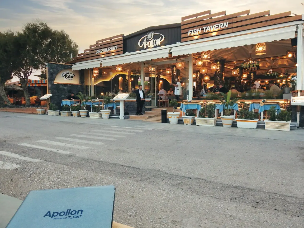 The lantern-lit terrace and sign of Apollon fish tavern on the Perissa beachfront at dusk, Santorini