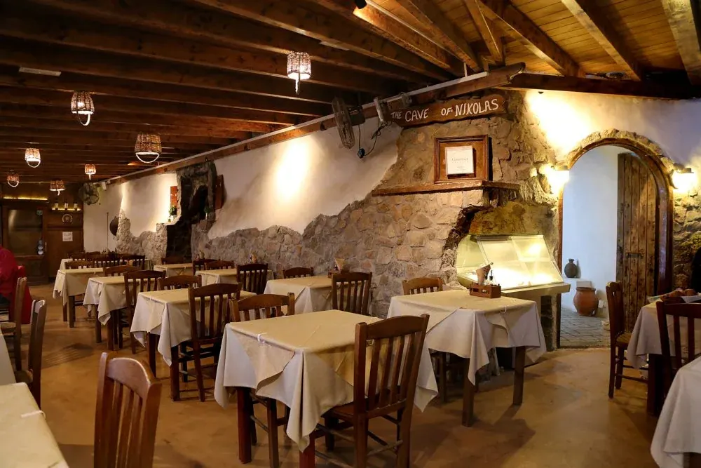 The stone-walled dining room of The Cave of Nikolas taverna, its name painted on a beam, at Akrotiri, Santorini