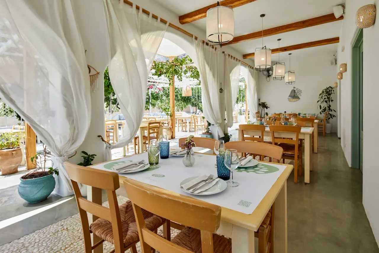 The bright whitewashed dining room and set tables of Fistikies restaurant, with white curtains opening to a courtyard, in Kamari, Santorini