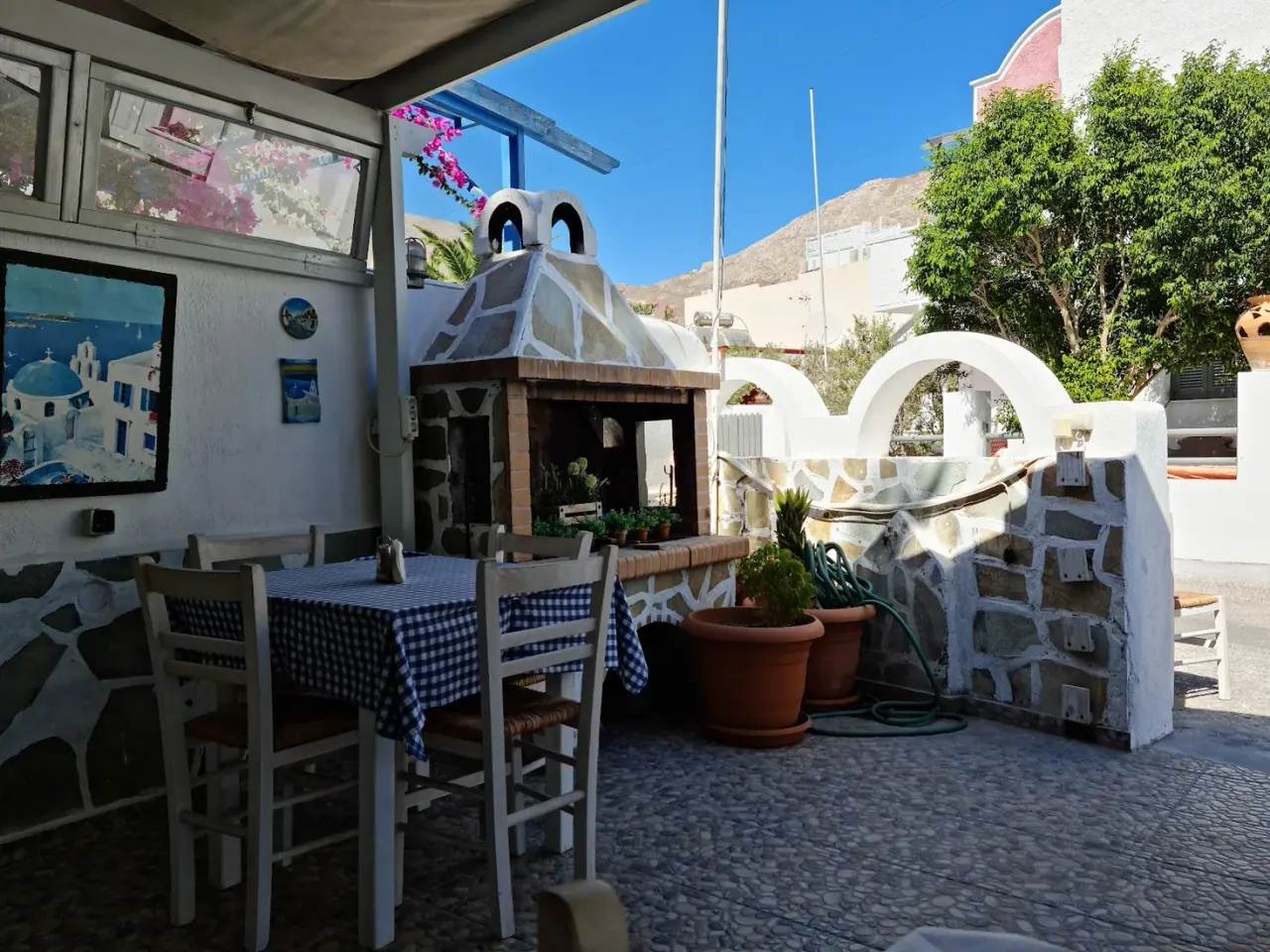 The stone-walled courtyard of Mama's Food taverna with a wood oven, blue-checked tablecloths and bougainvillea in Perissa, Santorini