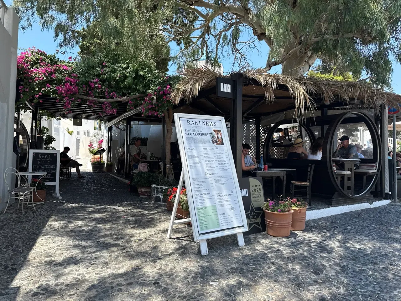 The thatched RAKI terrace with bougainvillea and a menu board on the square in Megalochori village, Santorini