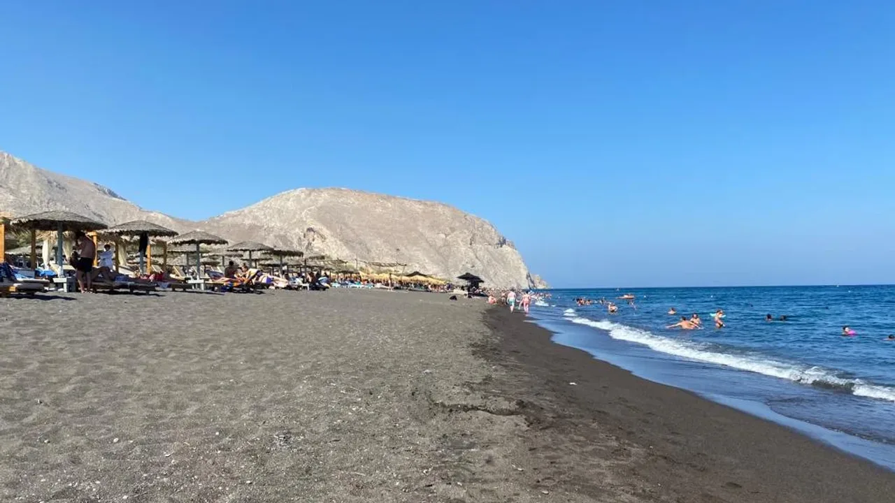 Straw umbrellas along the dark volcanic sand of Perivolos beach with swimmers in the sea below Mesa Vouno, Santorini