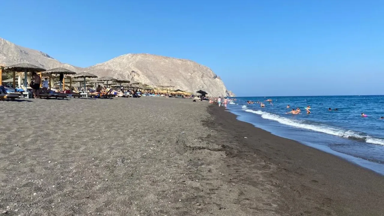 A black-sand beach day on Santorini with sunbeds and umbrellas by the calm sea