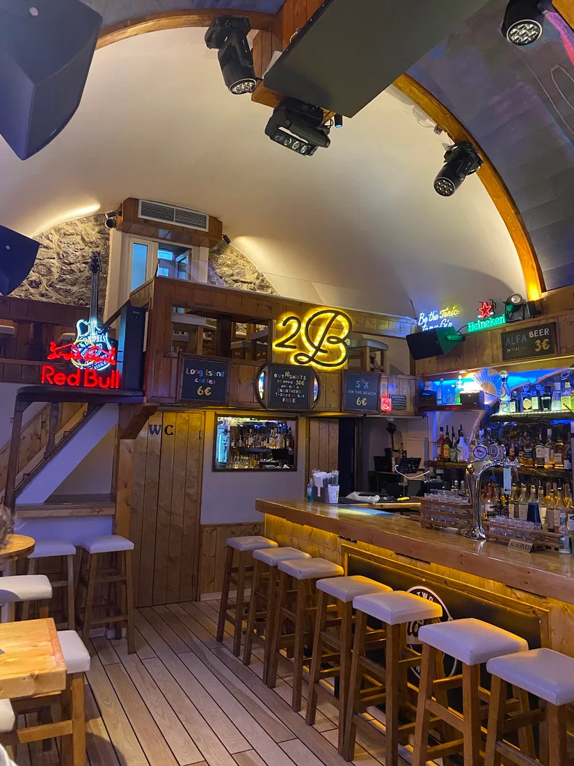 The wooden bar and stools under a vaulted ceiling with a glowing '2B' neon sign at 2 Brothers Bar, Fira