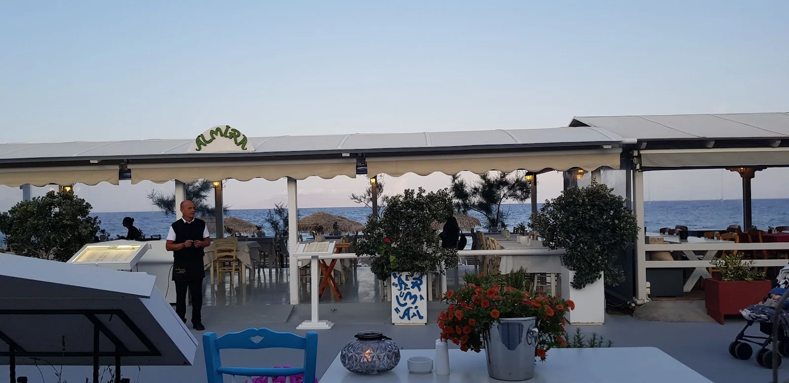 The awning-shaded ALMIRA seafront terrace looking out to the sea at dusk in Kamari, Santorini