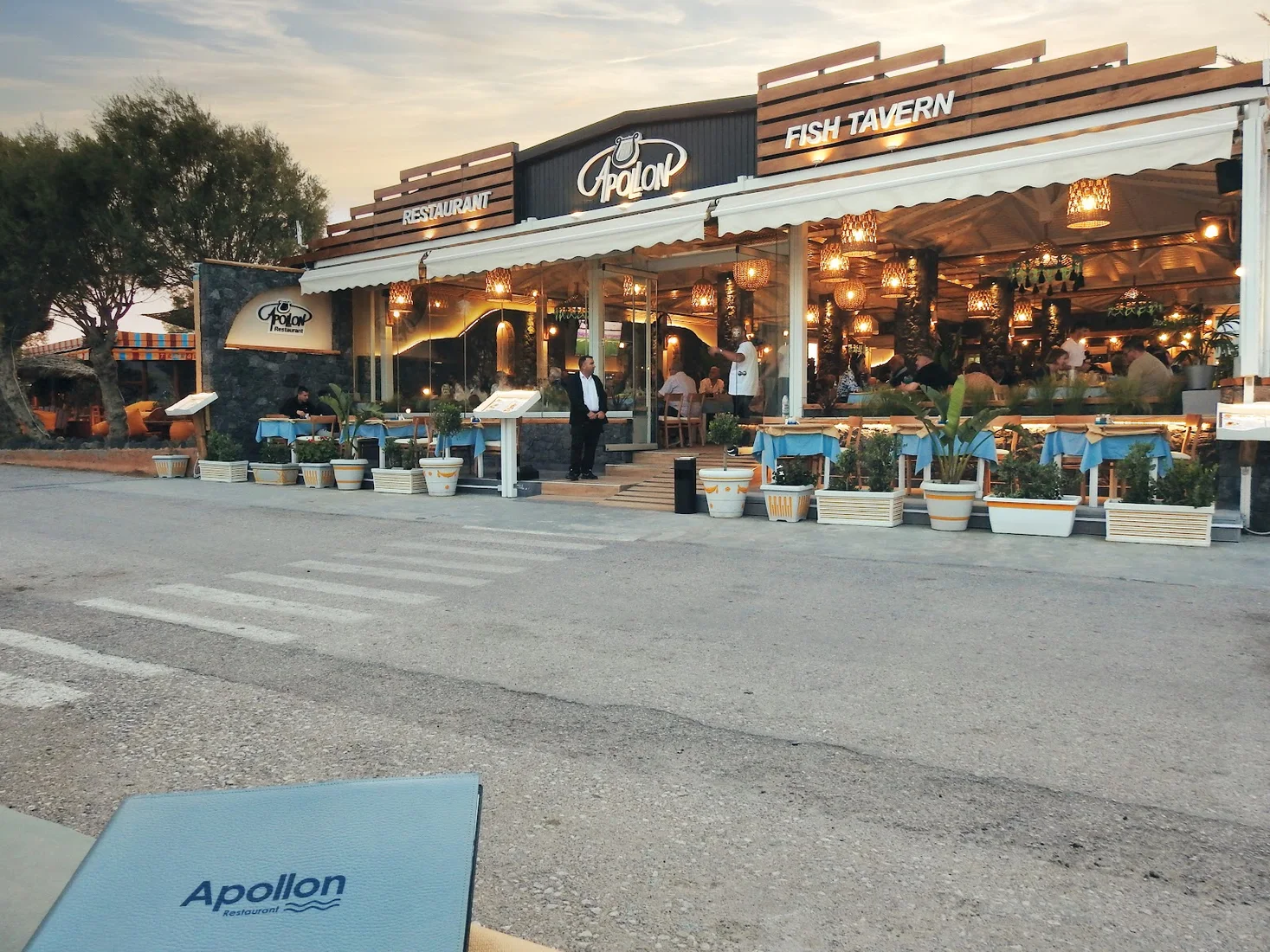 The lantern-lit terrace and sign of Apollon fish tavern on the Perissa beachfront at dusk, Santorini