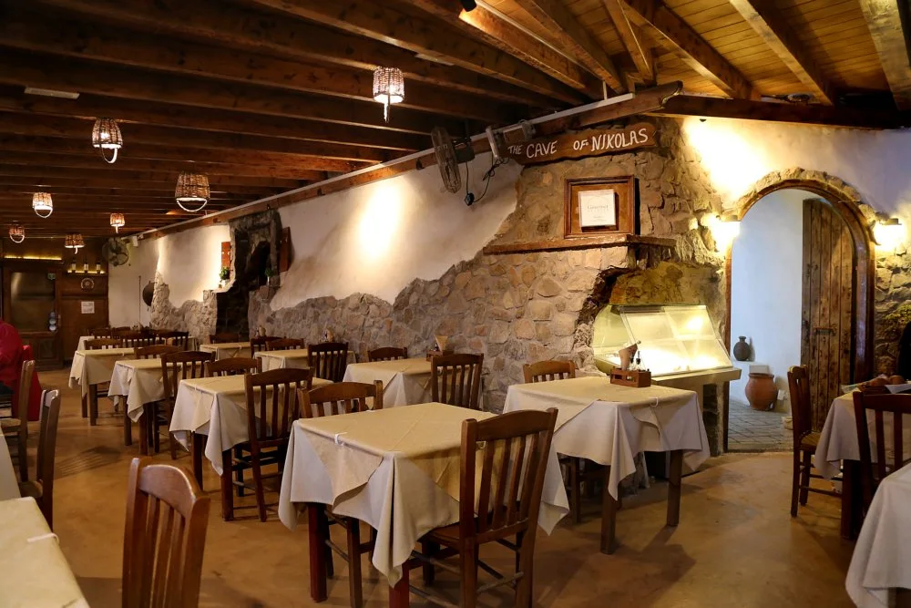 The stone-walled dining room of The Cave of Nikolas taverna, its name painted on a beam, at Akrotiri, Santorini