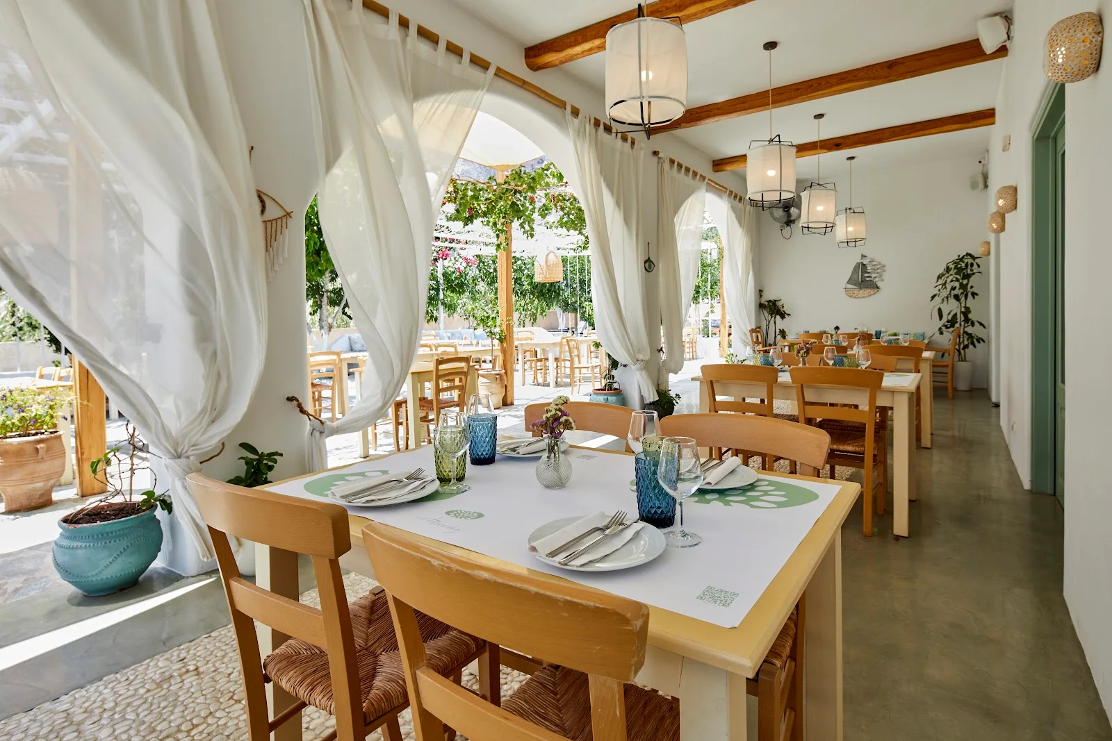 The bright whitewashed dining room and set tables of Fistikies restaurant, with white curtains opening to a courtyard, in Kamari, Santorini
