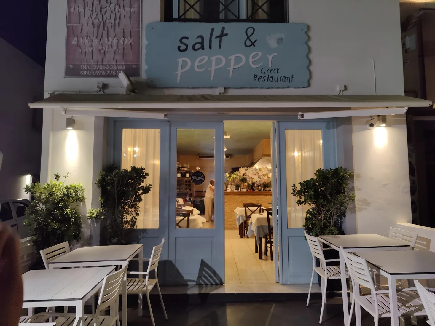 The lit blue-and-white facade and hand-painted sign of Salt & Pepper Greek restaurant in Fira, Santorini